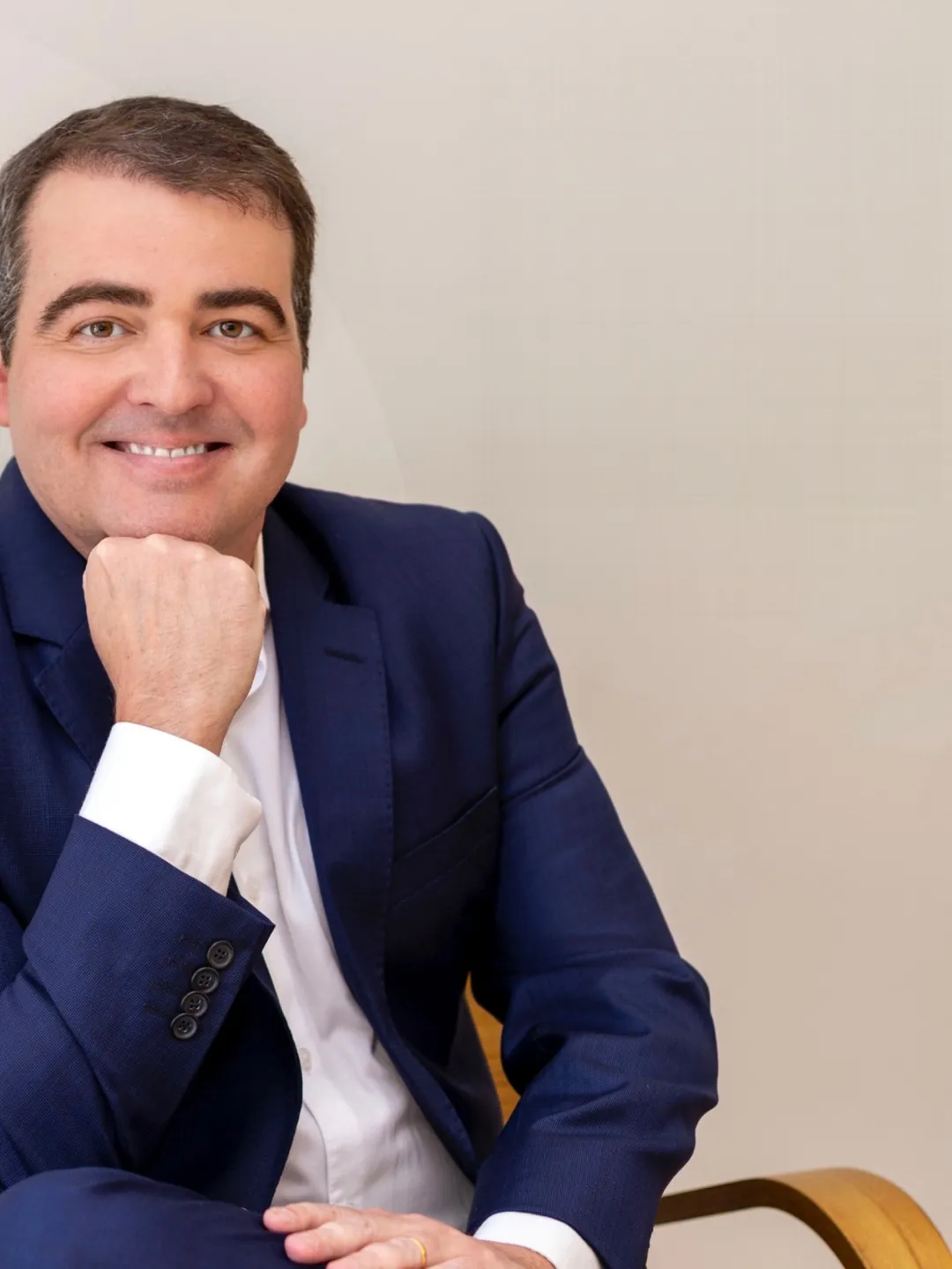 Dr. Diego Lima sentado, de terno azul-marinho, com a mão sob o queixo e um sorriso atento, diante de uma parede creme.