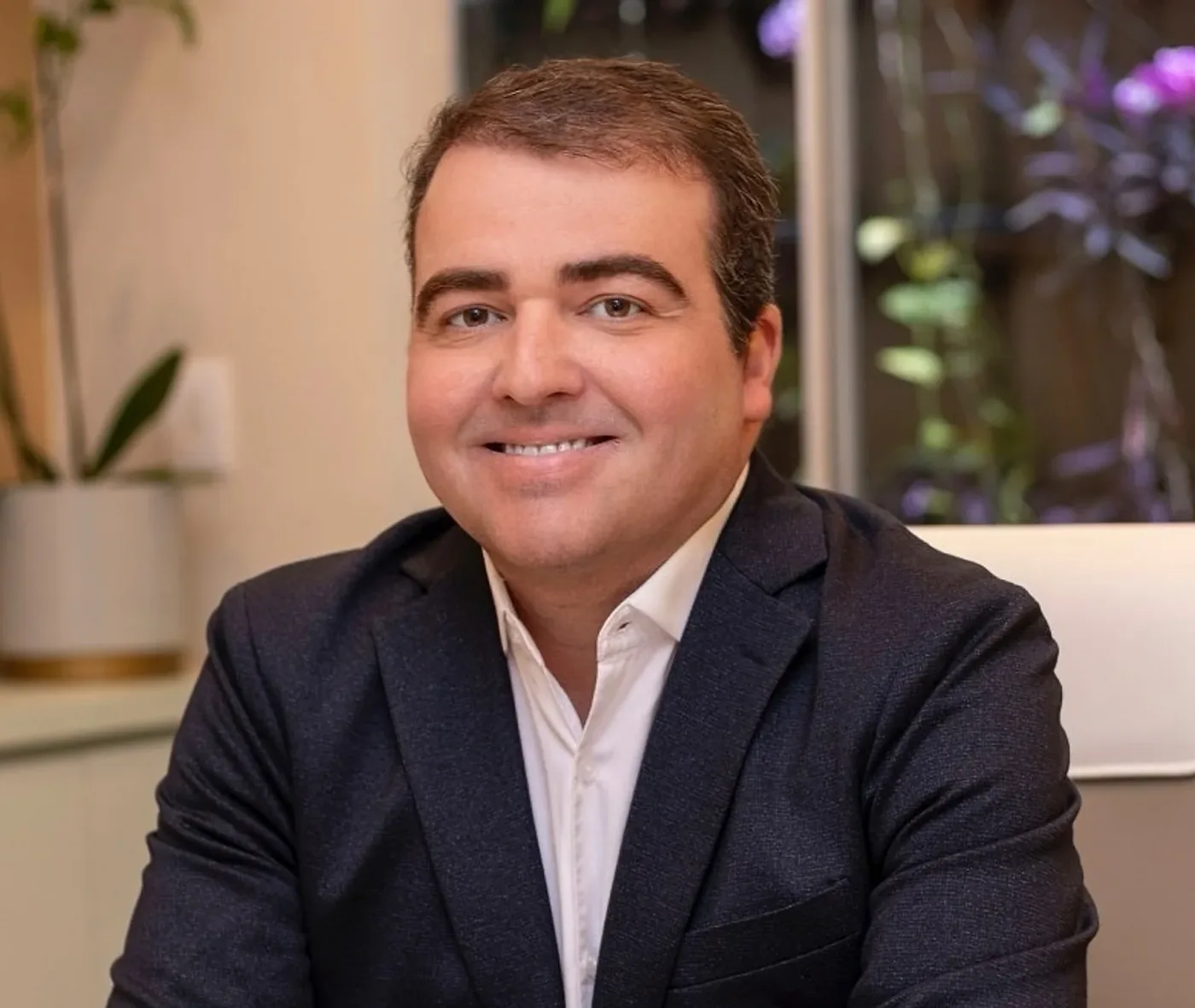 Dr. Diego Lima sorrindo, sentado à mesa do consultório, com o jardim vertical de orquídeas atrás do vidro