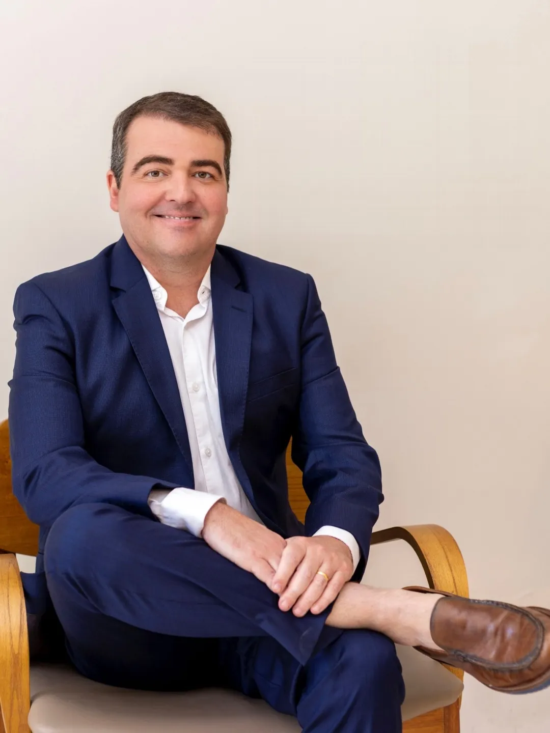 Dr. Diego Lima sentado numa cadeira de madeira, de terno azul-marinho e camisa branca, sorrindo, com as mãos sobre o joelho